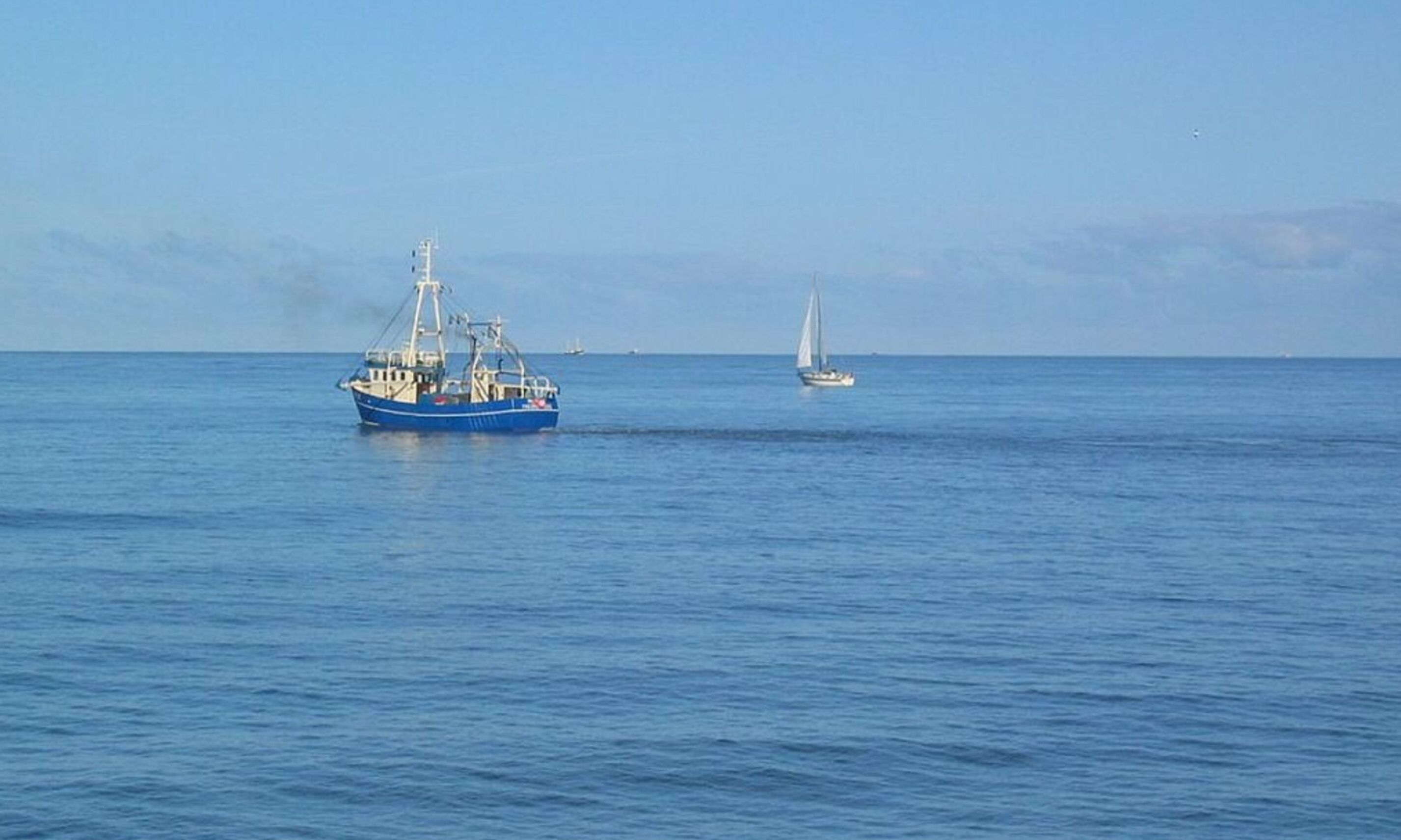 A blue fishing boat on calm waters with a sailboat in the distance.