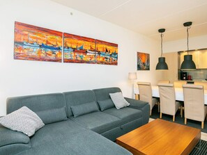 Una sala moderna con un sofá gris, una mesa de centro de madera y un comedor con sillas y una mesa. Hay dos cuadros grandes en la pared que representan una escena de atardecer.