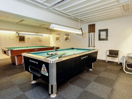 Ein Billardtisch mit schwarzem Tuch und Queue, ein weiterer Billardtisch im Hintergrund sowie ein Stuhl auf der rechten Seite.