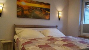 Quarto com cama de madeira, roupa de cama branca, colcha, duas luzes na parede e um quadro na parede.