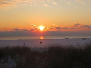 A beach scene with the sun setting over the horizon, a calm sea, and a few people walking along the shore. - "luv" in House Smaswai 10 (Nebel)
