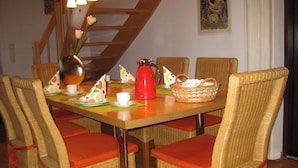 A dining area with wicker chairs and orange cushions, a wooden table with a red kettle, and a staircase with a wooden railing. - Holiday Home Haus Mewa (Neuenkirchen)