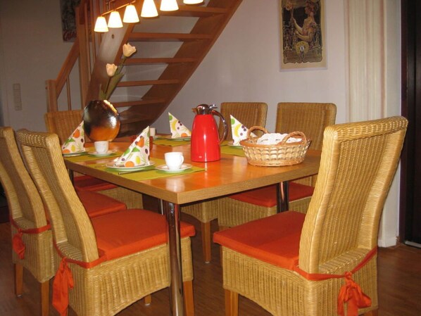 A dining area with wicker chairs and orange cushions, a wooden table with a red kettle, and a staircase with a wooden railing.