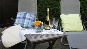 Two outdoor lounge chairs with cushions, a table with a bottle of champagne, glasses, and a vase with flowers. - Holiday Home Haus Mewa (Neuenkirchen)