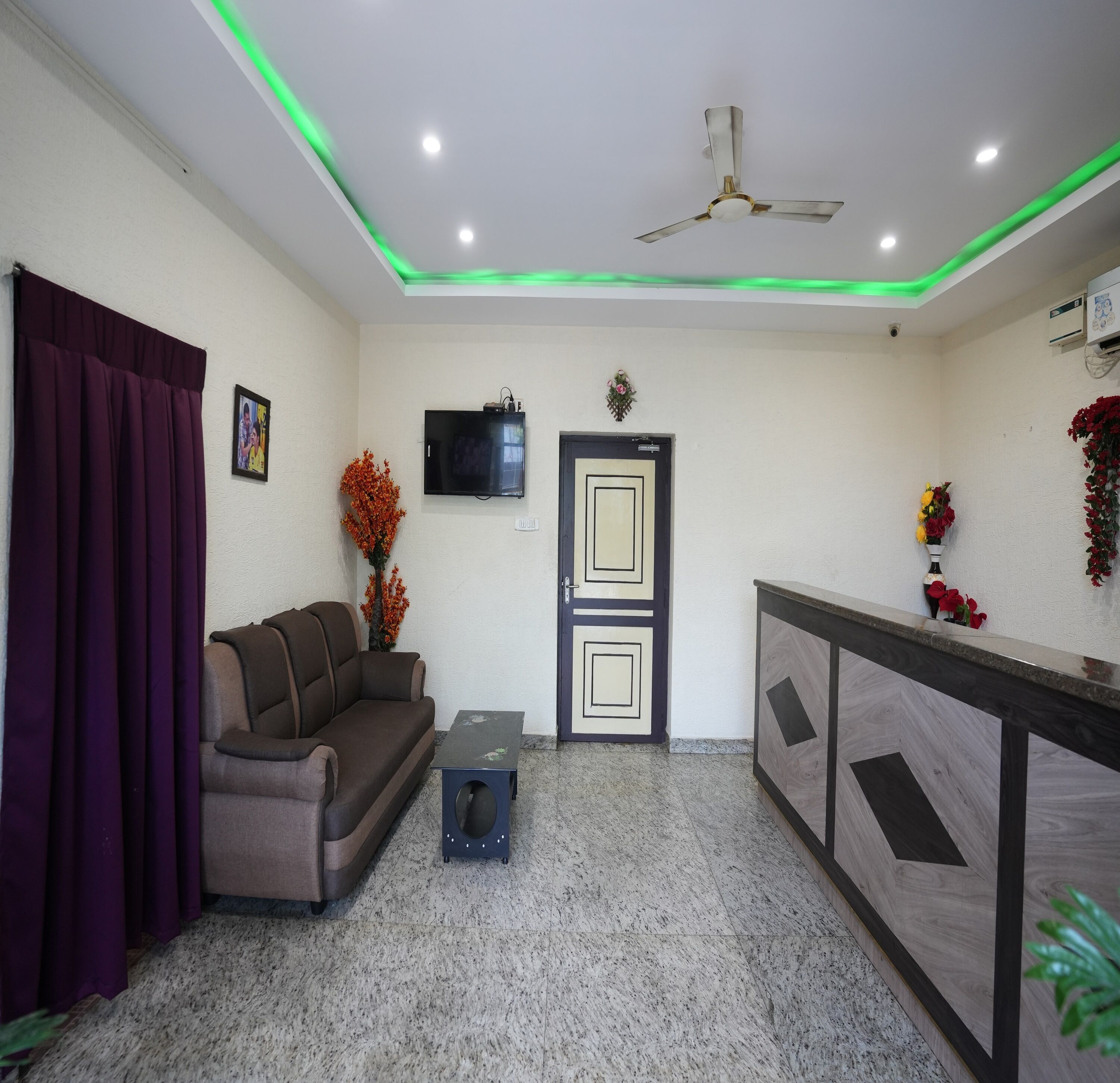 A reception area with a sofa, a small table, a TV, a door, and a ceiling fan.
