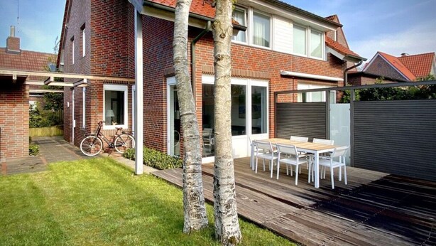 Una casa de dos pisos con techo de tejas rojas, fachada de ladrillo y terraza de madera con mobiliario exterior.
