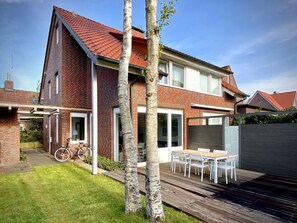 Una casa de dos pisos con techo de tejas rojas, fachada de ladrillo y terraza de madera con mobiliario exterior.