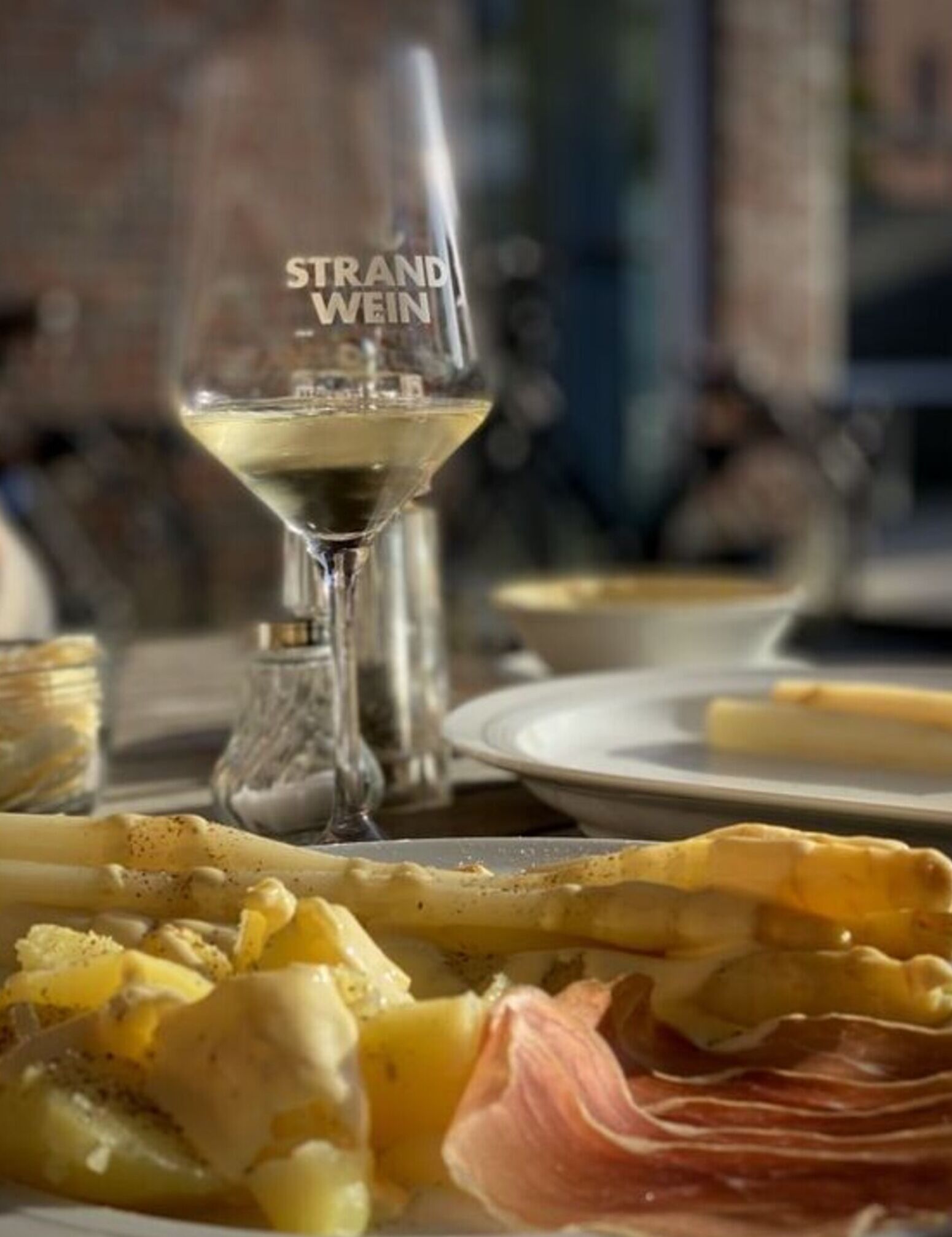 A glass of white wine with the label "STRAND WEIN" on a table with plates of food, including a dish with bacon and cheese.