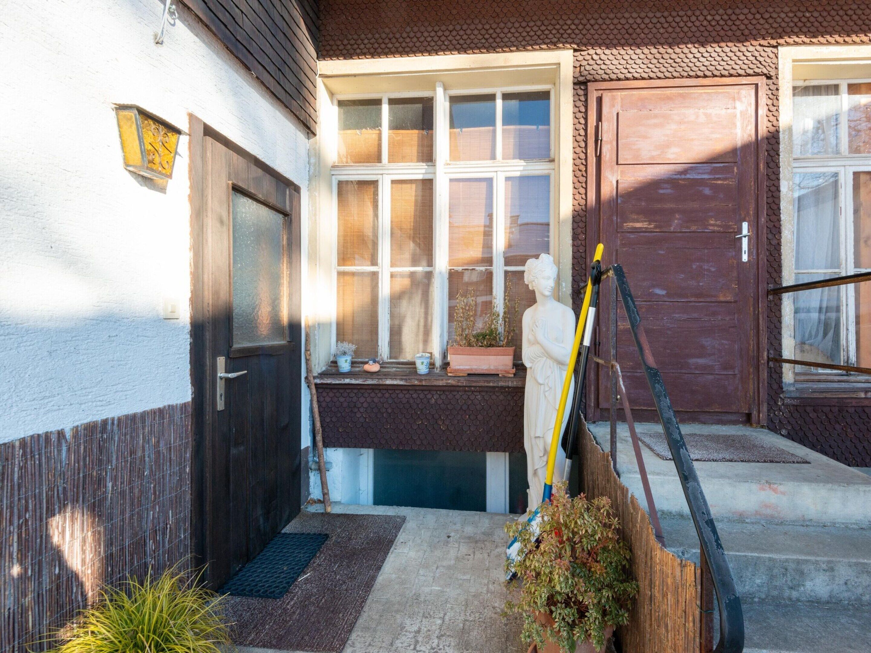 Une porte d’entrée en bois, une fenêtre avec des stores, un petit porche agrémenté d’un pot de fleurs et d’une statue.