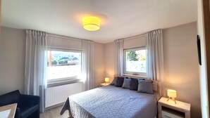 A bedroom with a bed, two bedside tables with lamps, a window with curtains, and a view of buildings outside.