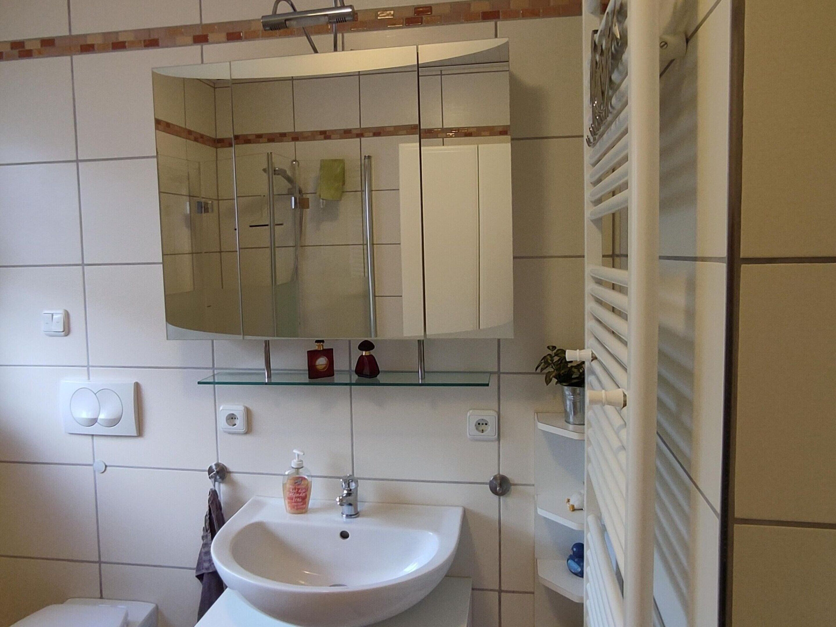 Ein Badezimmer mit einem weißen Waschbecken, einem Spiegel darüber und einem Glastischchen mit Toilettenartikeln.