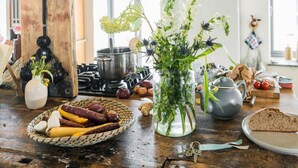 A kitchen counter with a variety of food items, a stove, a vase with flowers, and a window in the background. - Holidays in Happiness (Tetenbüll)