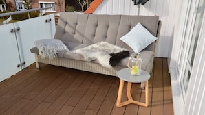 En træterrasse med en grå sofa, et lille rundt bord og et glas med et lys.