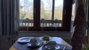 A dining table set with plates, cups, and cutlery, positioned near a window with a view of trees and a wooden deck. - Huis Hochfirst Appartement 20 (Titisee-Neustadt)