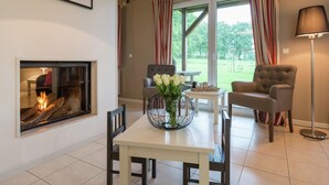 A living room with a fireplace, two armchairs, a dining table with a vase of flowers, and a view of the outdoors through large windows. - Luxury Holiday Home With Private Garden (Waimes)