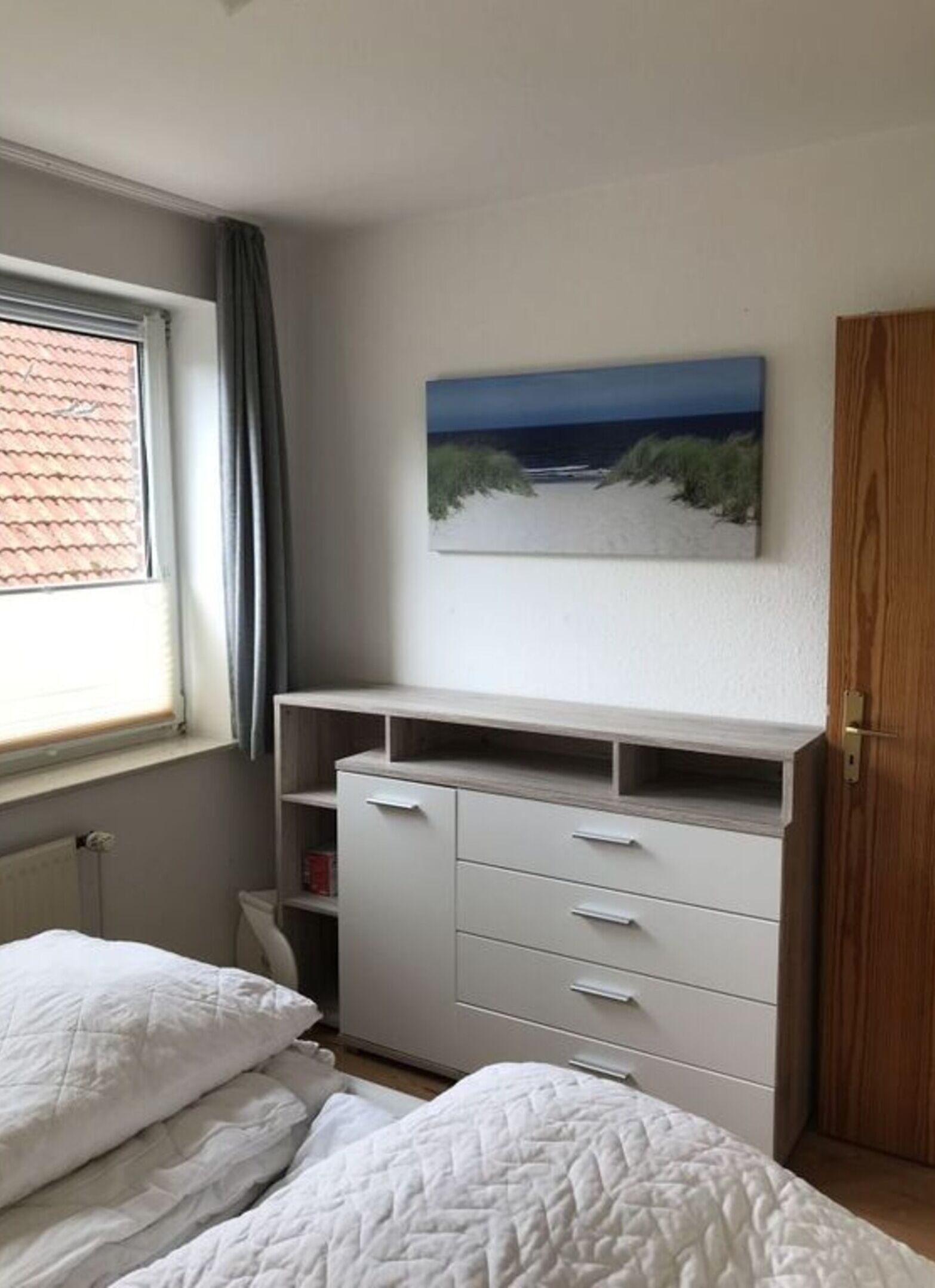 A bedroom with a bed, a dresser, a painting of a beach, and a window with curtains.