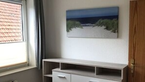 Une chambre à coucher avec un lit, une commode, un tableau représentant une plage et une fenêtre avec des rideaux.