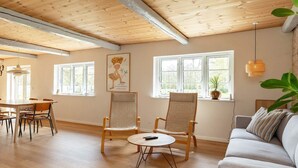 A spacious room with a high ceiling, wooden beams, a dining area with a table and chairs, a sofa, and large windows allowing natural light. - 5 Star Holiday Home in Laeso-by Traum (Læsø)