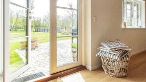 A room with a large sliding glass door leading to a patio with potted plants and a wooden fence. There is a wicker basket with cushions and a small table with a vase. - 5 Star Holiday Home in Laeso-by Traum (Læsø)