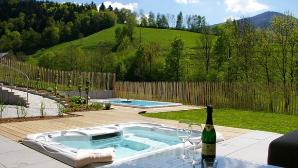 A backyard with two hot tubs, a wooden deck, and a glass table with wine glasses and a bottle. - Tasteful Apartment in Elzach-yach (Elzach)