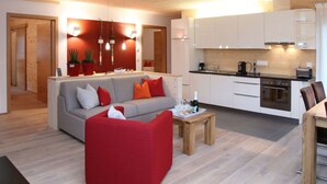 A modern living room with a sofa and a red armchair, a wooden coffee table, and a kitchen area with white cabinets and a stove. - Tasteful Apartment in Elzach-yach (Elzach)