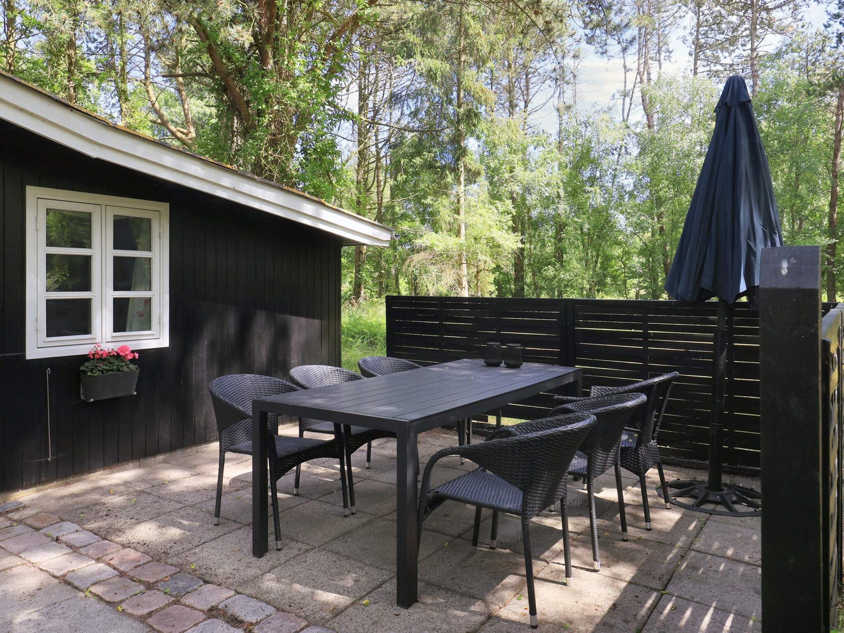 Eine Terrasse mit Tisch und Stühlen, ein kleines Gartenhaus mit Fenster und ein dunkel gefärbter Sonnenschirm.
