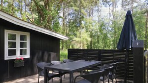 Une terrasse avec une table et des chaises, un petit abri de jardin avec une fenêtre et un parasol de couleur foncée.
