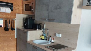 Cocina compacta con armarios grises, un lavamanos blanco y un microondas. Las paredes son de madera y hay una pequeña encimera.