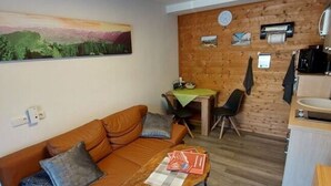 Un espacio de sala acogedor con un sofá de cuero marrón, una mesa de centro de madera y una mesa de comedor con sillas. Se ve un área de cocina al fondo, con armarios de madera y un fregadero.