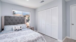 Une chambre à coucher avec une tête de lit capitonnée, une grande fenêtre, une armoire et une table de chevet avec une lampe.