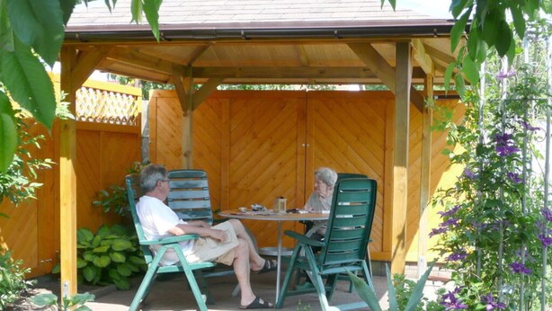 A covered patio with two people seated at a table, surrounded by wooden fencing and potted plants. - Ostenberg - Near CJD Comfortable Holiday Residence (Garz/Rügen)