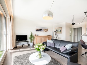 En moderne stue med en sort lædersofa, et hvidt sofabord og et fjernsyn. Der er en plante på gulvet og et billede på væggen.