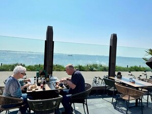Un comedor con vista al mar, con mobiliario exterior y una mesa con comida y bebidas.