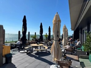 An outdoor patio with round tables and chairs, umbrellas, and a view of the sea. - Stay in Scherpenisse Near Oosterschelde (Scherpenisse)