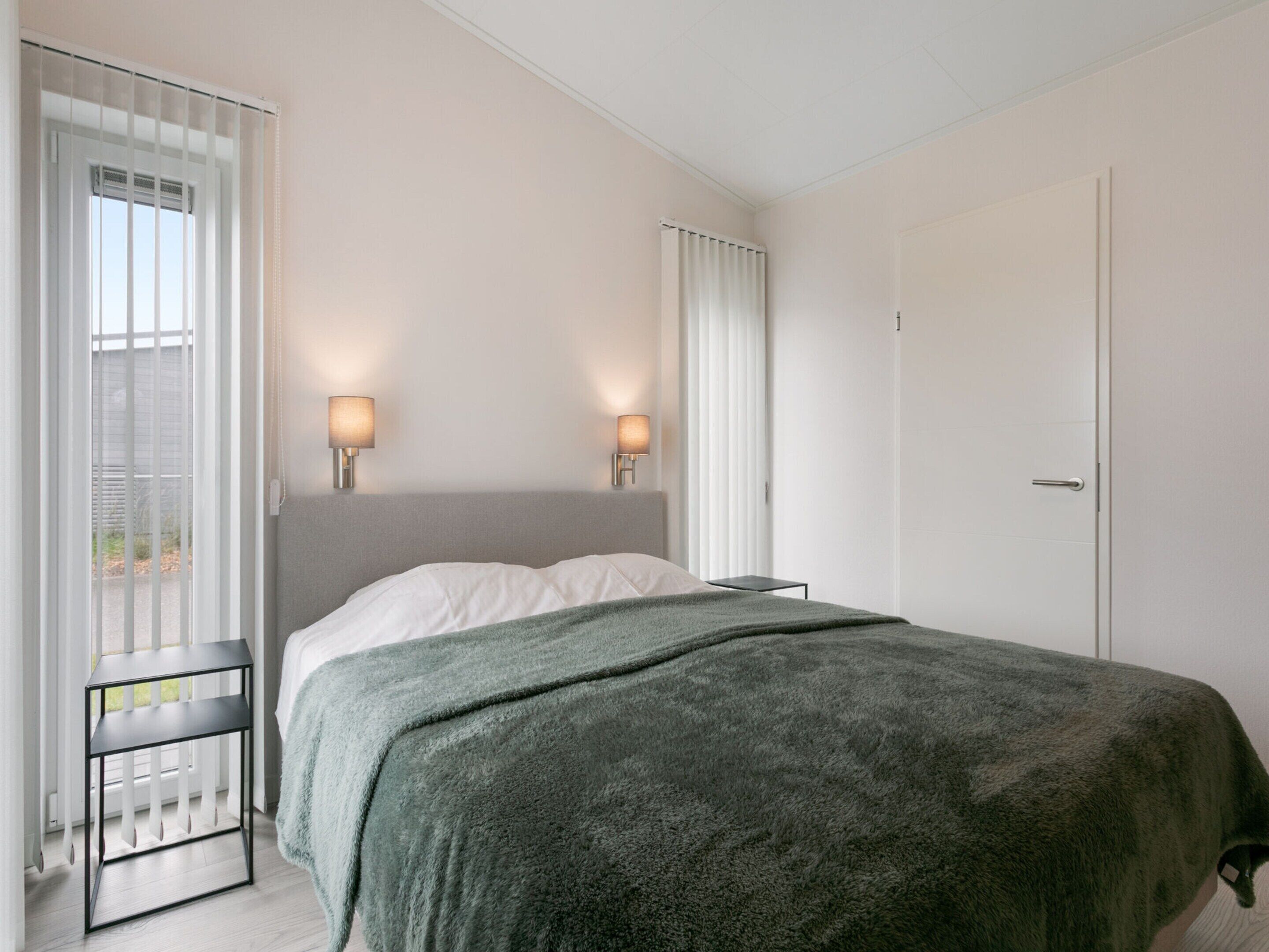 A bedroom with a bed, two bedside lamps, a window with blinds, and a door.