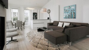 Uma sala de estar moderna com um sofá modular cinza, uma mesa de centro e um tapete felpudo. Há uma área de jantar com uma mesa e cadeiras brancas, e uma cozinha com armários brancos e uma luminária pendente.