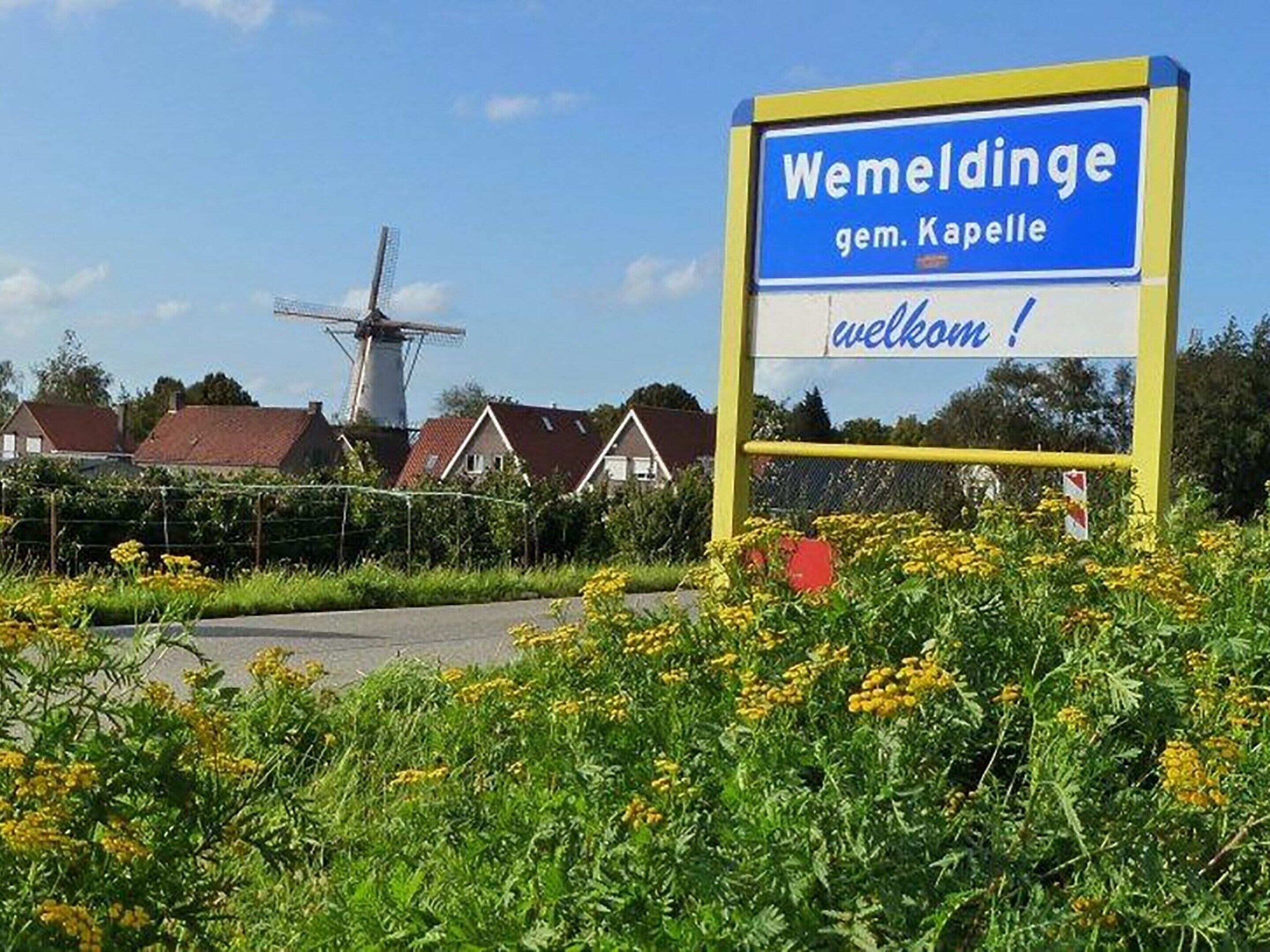 Señal de bienvenida a Wemdinge, un pueblo en Alemania, con un molino de viento y casas al fondo.