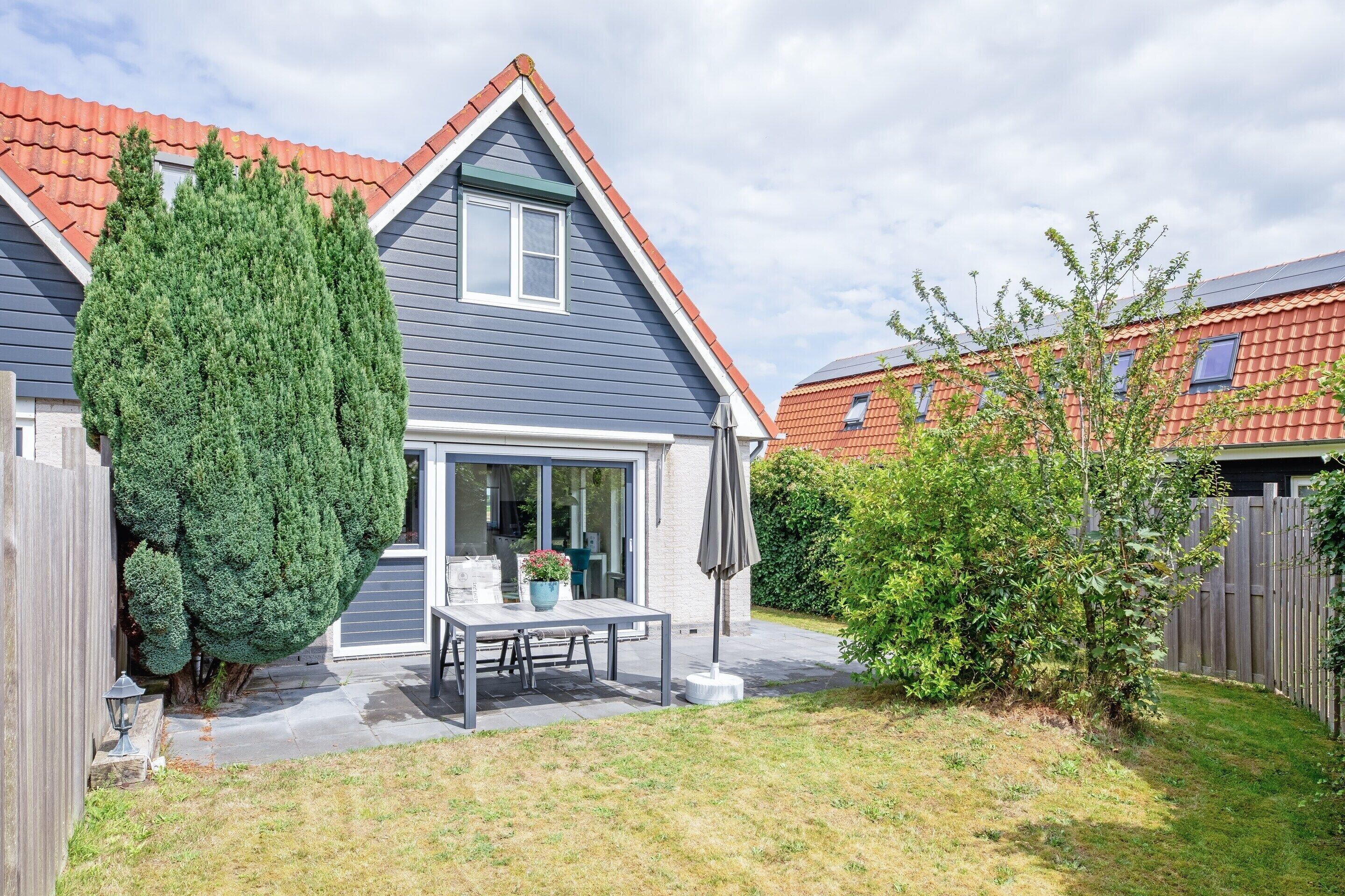 A modern house with a grey roof, a large coniferous tree, and a patio with a table and chairs.