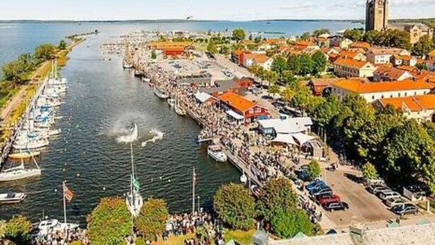 Un pueblo a orillas de un río con una imponente torre de iglesia, un puerto deportivo con barcos, y una calle con edificios muy cerca unos de otros.