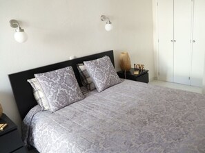 A neatly made bed with patterned pillows, a bedside table with a lamp, and a wardrobe in a simple room. - Apartment on the Beach in Vale Centianes (Lagoa)