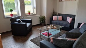 A modern living room with a glass coffee table, two sofas, a small side table, and a window with a view of greenery. - Holiday Apartment With a Terrace (Bremerhaven)