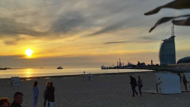 A beach scene with a sunset, a prominent building, and people walking. - Holiday Apartment With a Terrace (Bremerhaven)