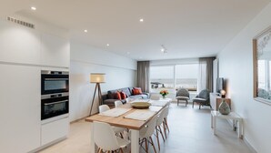 Una sala de estar moderna y bien iluminada con comedor, un conjunto de sofás y vista al mar.