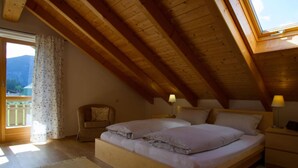 A bedroom with a wooden ceiling, a bed with pillows, two bedside tables with lamps, a chair, and a view of mountains through a window. - Lovely Apartment in Oberammergau (Oberammergau)
