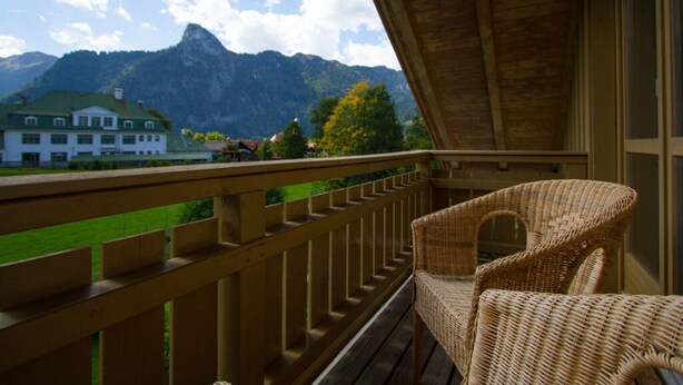 A balcony with a wicker chair, wooden railing, and a view of mountains and a white building. - Lovely Apartment in Oberammergau (Oberammergau)