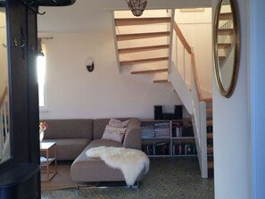 A cozy living room with a sofa, a white furry ottoman, a bookshelf, and a staircase.