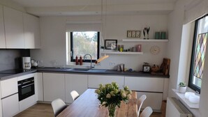 A modern kitchen with white cabinetry, a wooden dining table, and a window with a view of trees. - Villa Right by the sea (Julianadorp)