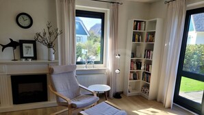 A modern living room with a fireplace, a comfortable chair, a small table, a bookshelf, and a large window with a view of the outdoors. - Villa Right by the sea (Julianadorp)