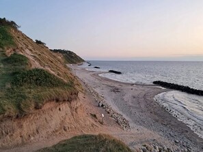 Paisagem costeira com uma praia de areia, dunas gramadas e um afloramento rochoso.