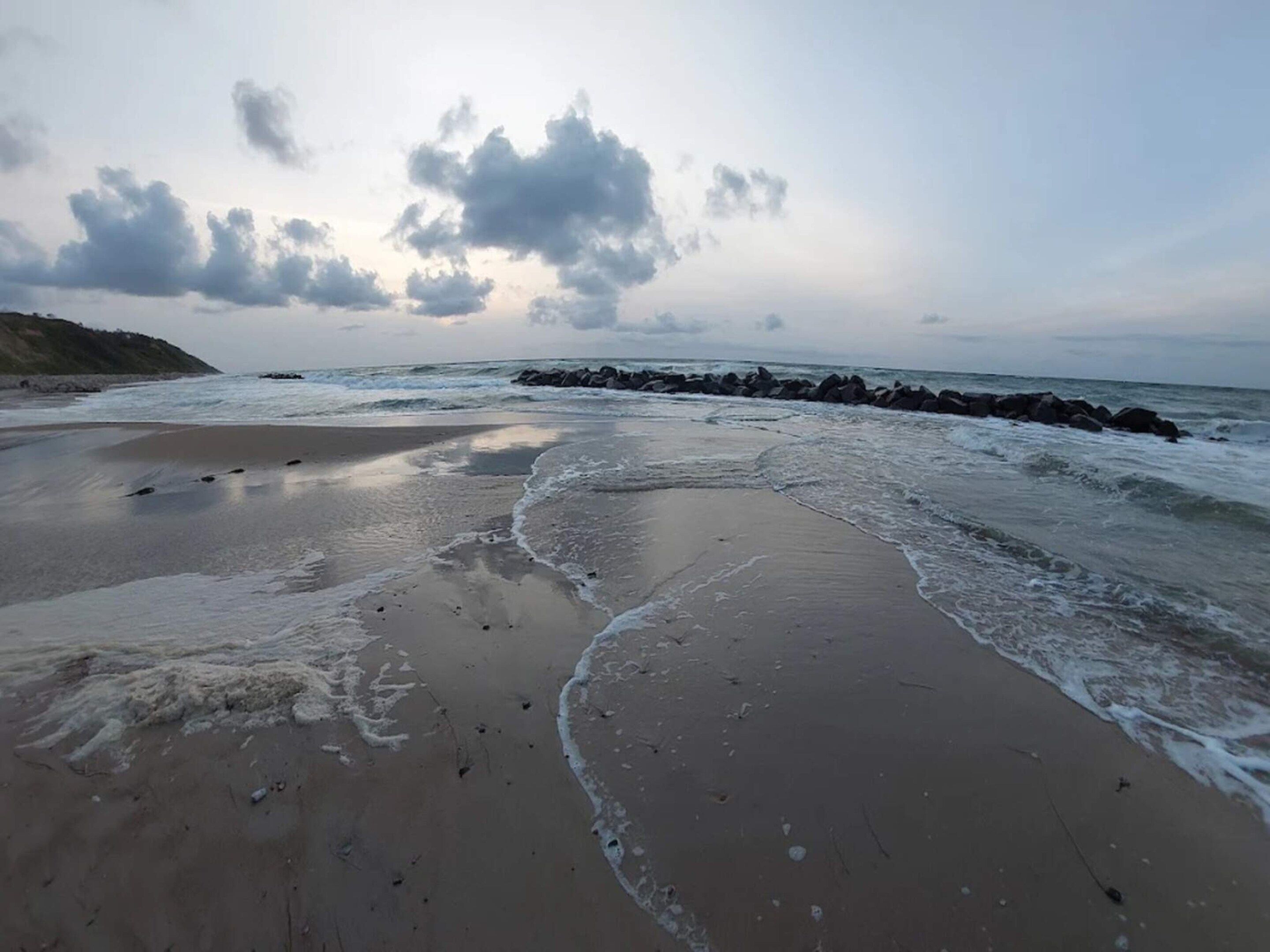 A beach with waves, a rocky breakwater, and a cloudy sky.